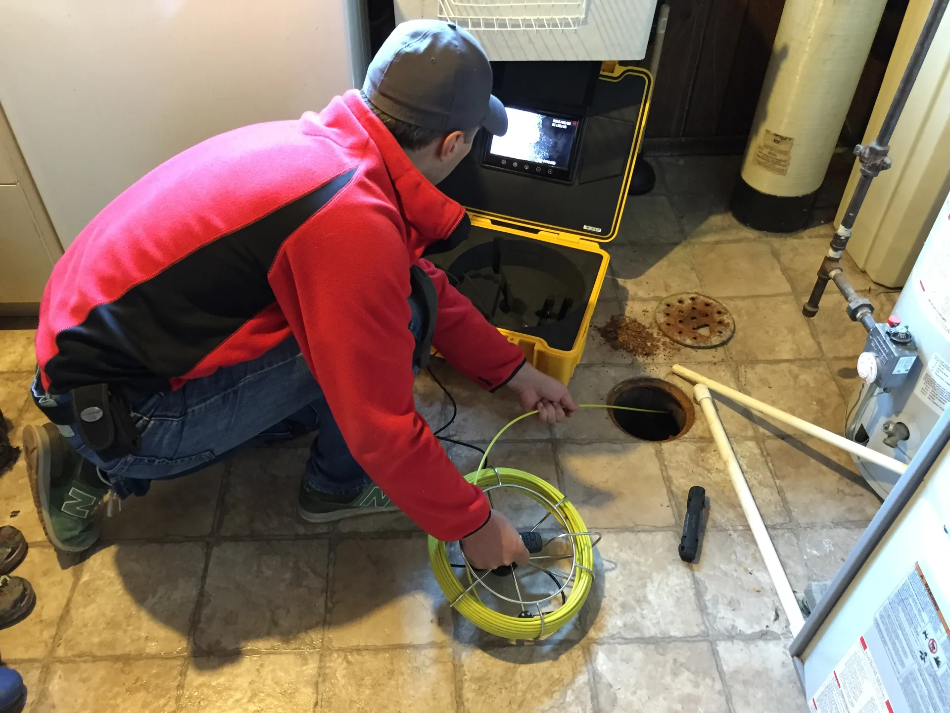 A technician feeding a drain camera into a basement cleanout while the line shows on a monitor